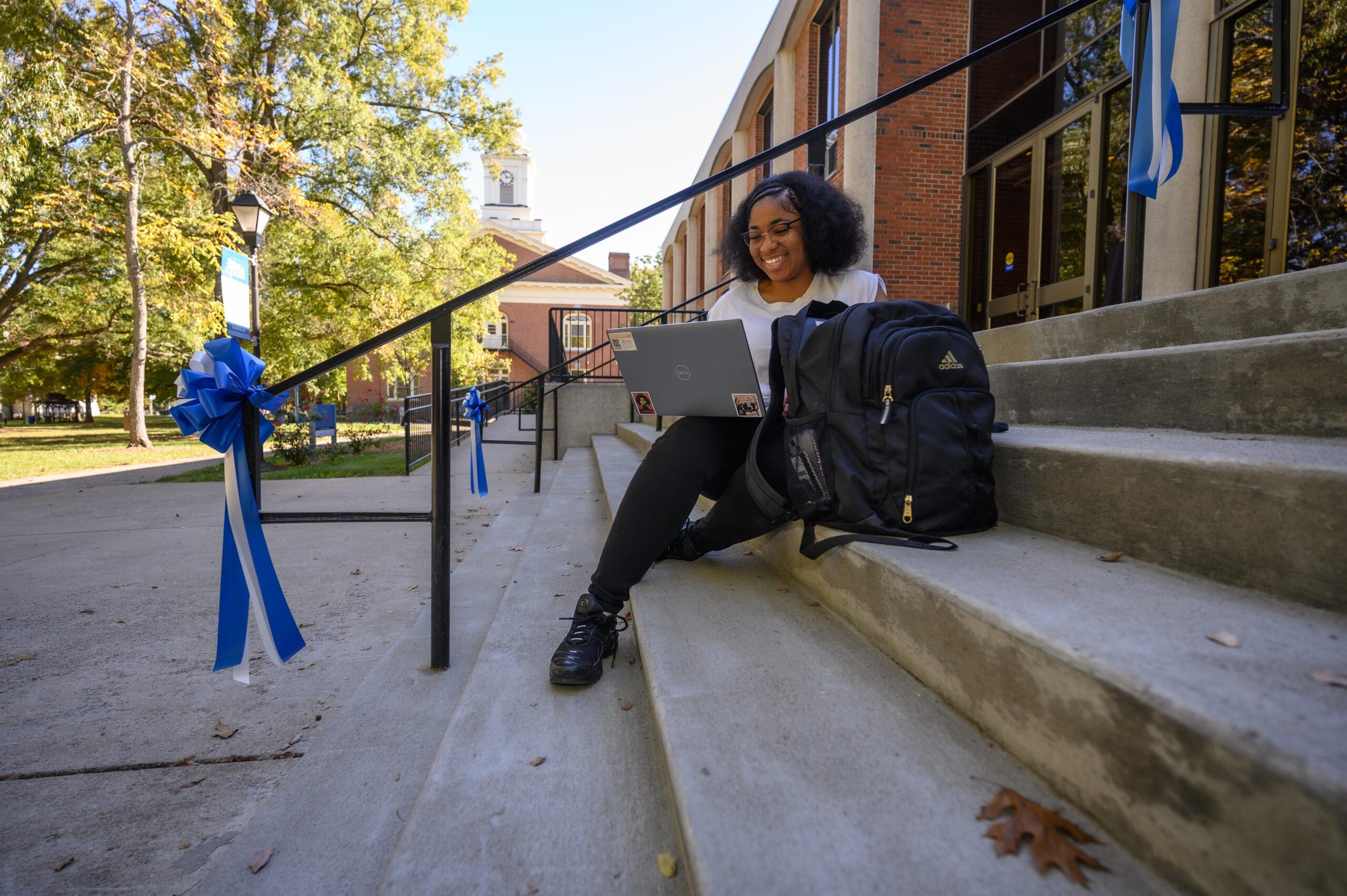 A female student sits on the stairs in front of Hutchins Library, smiling at her computer.