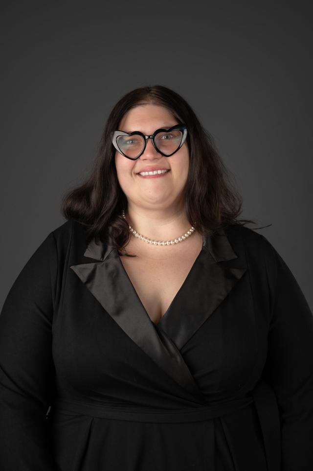 Sam Cole portrait wearing a black dress with black glasses smiling and behind is a grey background