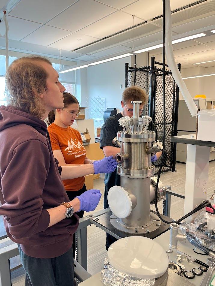 Three Berea College physics students work together on a machine in a lab classroom.