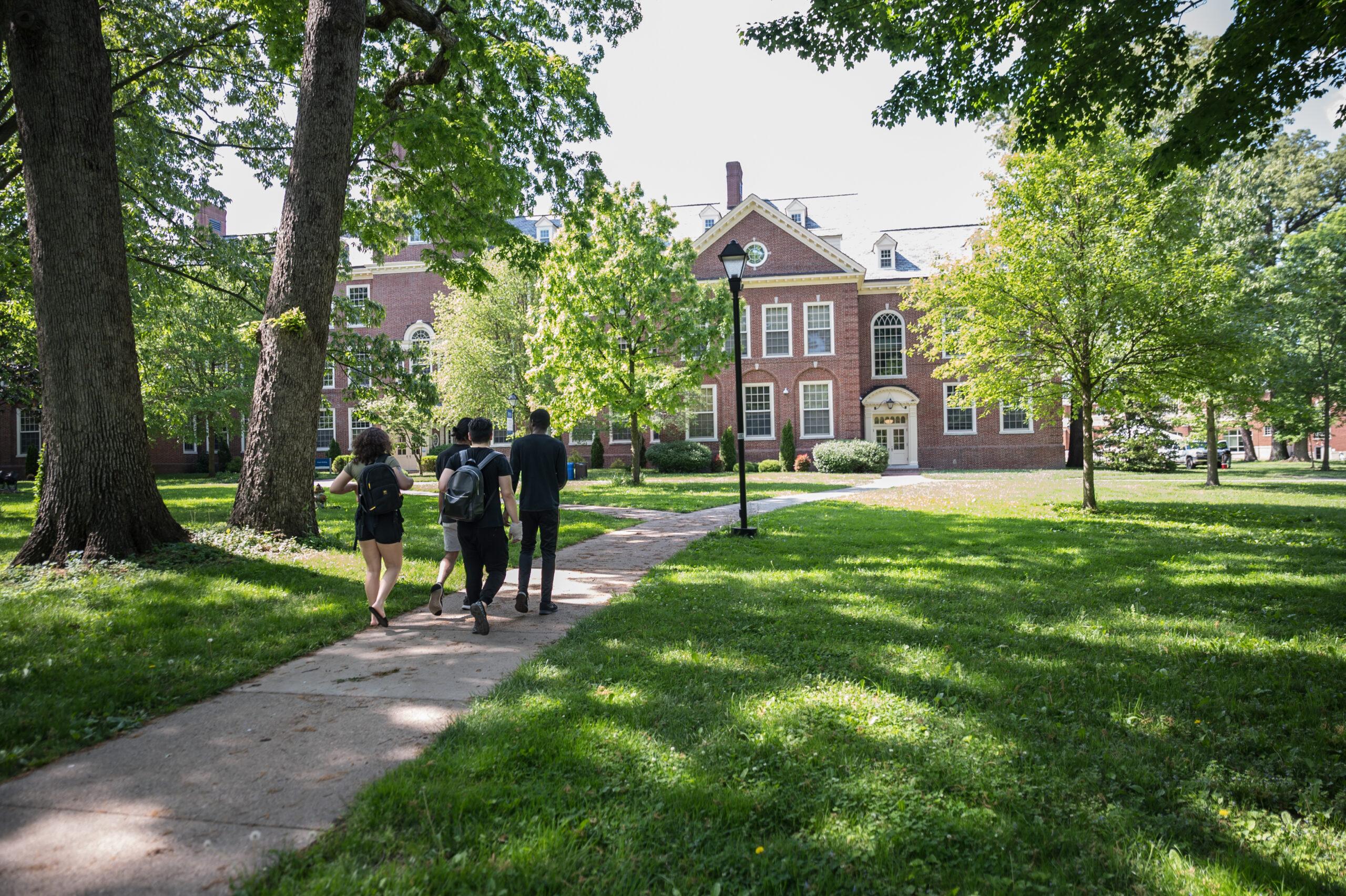 A photo of a very green campus with four students walking away from the camera, with their faces facing it, towards a brick building.